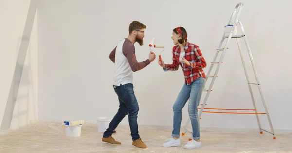 Młoda wesoła para poruszająca się zabawnie przed kamerą podczas naprawy w swoim nowym mieszkaniu przed zamieszkaniem razem. Wewnątrz — Zdjęcie stockowe