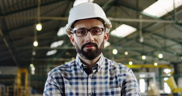 Close-up van een fabrieksarbeider in een helm, bril en ruitshirt poserend voor de camera in een groot leeg fabriekshuis. Naar binnen. Portretfoto — Stockfoto