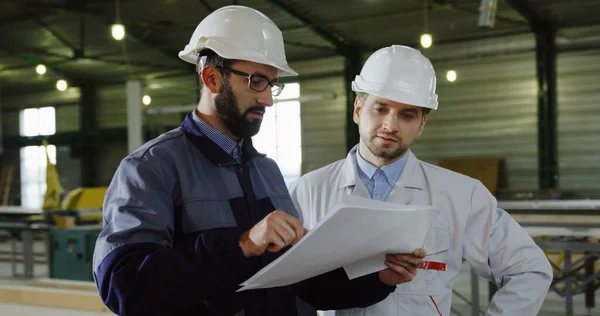 Mannelijke werknemer van de fabrikant en zijn ingenieur in helmen bespreken het project dat ze zien op de grote papieren tekeningen. Fabriekskamer. Binnenshuis. Portretfoto. — Stockfoto