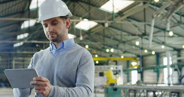 Portret van aantrekkelijke mannelijke ingenieur in een helm tape en scrollen op zijn tablet, dan kijken in de camera en glimlachen in de grote industriële faciliteit. Binnen. Sluiten. — Stockfoto