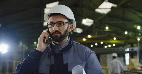 Blanke fabrieksarbeider met een helm in gesprek op de smartphone in het midden van de grote industriële kamer. Binnenshuis. Portret. Sluiten. — Stockfoto