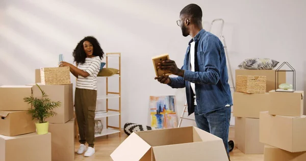 Handsome young African American man unpacking the carton box and showing a photo in the frame to his pretty curly girlfriend who standing behind. Couple moving in the new apartment. Indoor Stock Photo