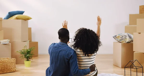 Parte trasera de la pareja afroamericana casada sentada en el suelo entre cajas y mirando el plano, considerando un diseño para su nuevo hogar. Adentro. Imagen de archivo