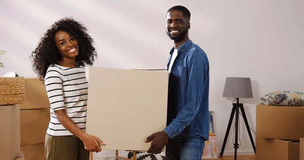 Happy young African American couple carrying a big carton box while moving in the new apartment and posing with it to the camera. Portrait shot. Indoor Stock Photo