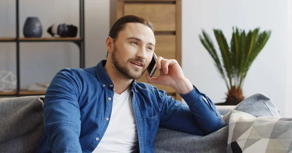 Handsome caucasian man sitting in the living room on the sofa and having a nice talk on the phone. Cozy interior of the home. Indoors Royalty Free Stock Images