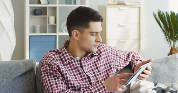 Young handsome man in the plaid shirt scrolling on the tablet device, watching something and laughing on the couch at home. Indoor Stock Image