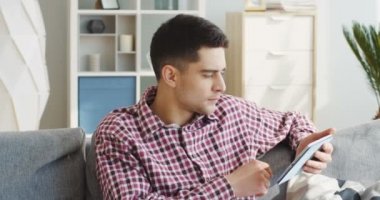 Ekose gömlekli genç yakışıklı adam tablet cihazına kayıyor, bir şey izliyor ve evdeki kanepede gülüyor. Kapalı