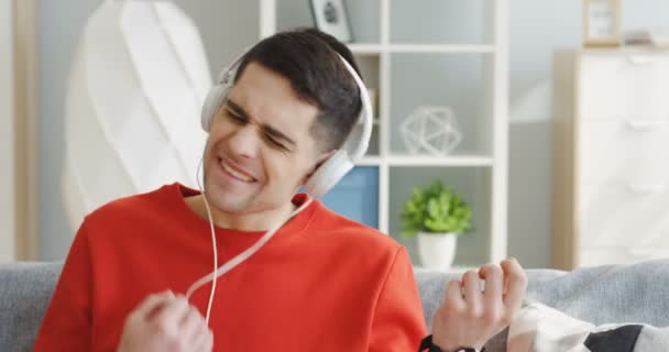 Portrétní záběr pohledného mladíka ve sluchátkách, jak poslouchá hudbu, vychutnává a napodobuje kytaru hrající na gauči v obývacím pokoji. Zavřít. Indoors — Stock video