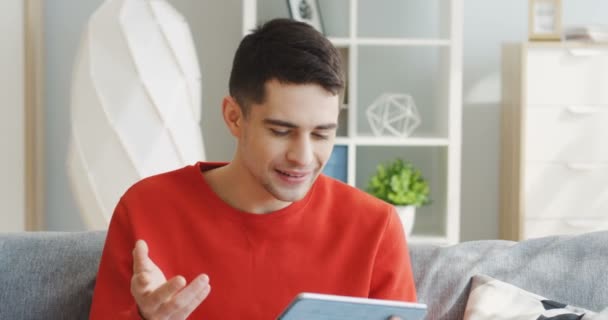 Caucásico joven hombre guapo en el suéter rojo tener un videochat mientras está sentado en el sofá con almohadas en casa. En interiores — Vídeos de Stock