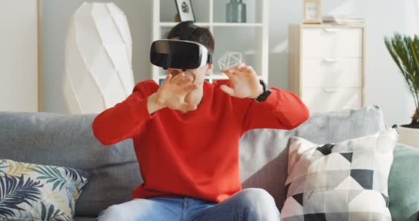 Porträtt av den unge kaukasiske mannen i röd tröja med VR-headset i VR-glasögonen på soffan med kuddar i det fina ljusrummet. Inomhus — Stockvideo