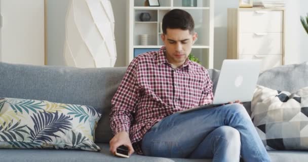 Jóképű fiatalember kockás ingben, aki a laptop számítógépén dolgozik, és telefonon beszél a nappaliban. Belül. — Stock videók