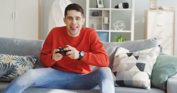 Emocionado joven caucásico hombre jugando videojuego en la sala de estar en el sofá y se mueve divertido. En interiores — Vídeos de Stock