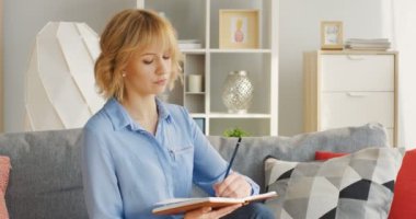 Sarışın, genç ve çekici bir kadın kanepede yastıklarla oturuyor ve rahat oturma odasındaki deftere veya günlüğe notlar yazıyor. Kapalı