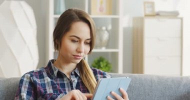 Gri koltukta oturan ve rahat odadaki tablet bilgisayarı kullanan güzel kadının portresi. İçeride.