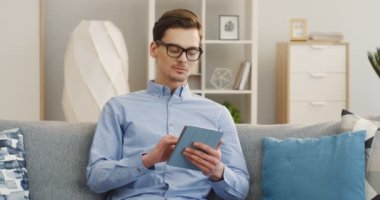 Gözlüklü genç çekici adam gri koltukta oturuyor ve tablet cihazını kullanıyor, ciddi bir şey düşünüyor. - Evde. Kapalı