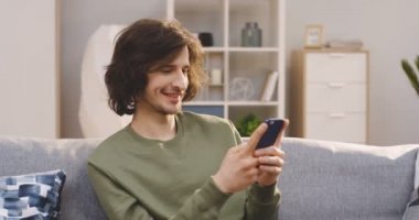 Evdeki kanepede otururken uzun saçlı, akıllı telefonuyla kayan ve bantlayan genç yakışıklı adam. İçeride.