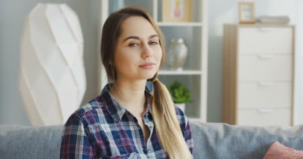 Ung vacker kvinna sitter på soffan och tittar på sidan och sedan vrider huvudet mot kameran och ler. Porträtt skjuten. Inomhus — Stockvideo