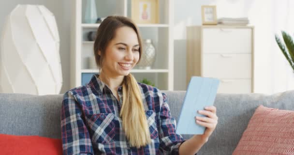 Mulher jovem caucasiana na camisa xadrez ter um videochat enquanto sentado no sofá cinza com almofadas em casa. Interior — Vídeo de Stock