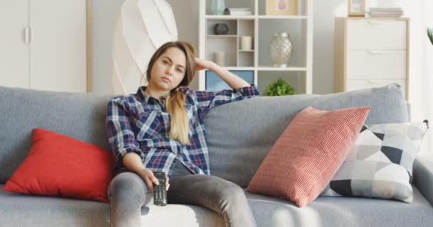 Młoda znudzona kobieta siedząca na kanapie, oglądająca telewizję i zmieniająca kanały z pilotem w ręku. W domu. Wewnątrz — Wideo stockowe