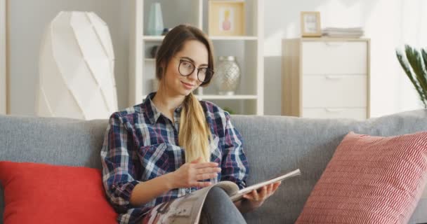Ung vacker kvinna i brokig blå skjorta videochattar medan du sitter på den grå soffan med kuddar hemma. Inuti — Stockvideo