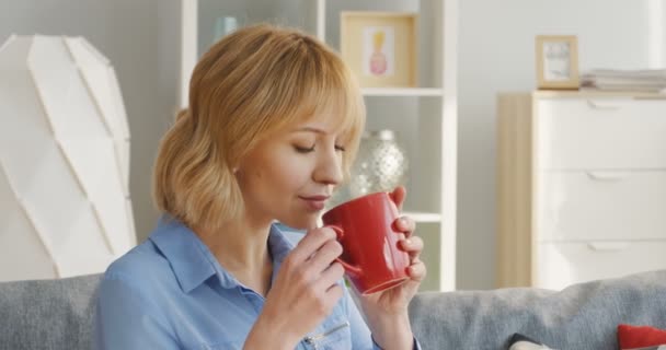 Retrato de la joven rubia hermosa mujer bebiendo té caliente o café de una taza roja y soñando mientras está sentada en el sofá en casa. Adentro. — Vídeo de stock