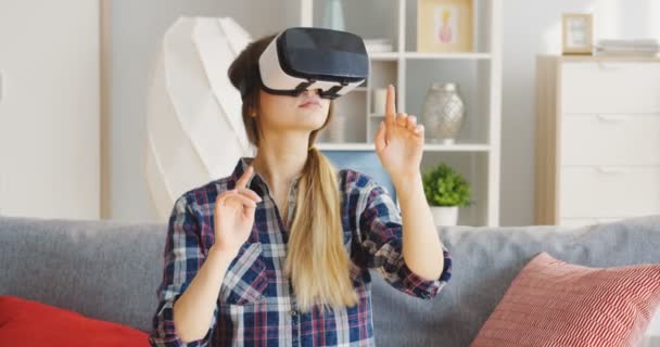 Retrato de la joven mujer caucásica vestida con una camisa a cuadros con auriculares VR en las gafas VR en el sofá con almohadas en la bonita sala de luz. En interiores — Vídeos de Stock