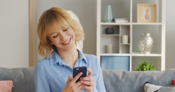 Ung glad blond kvinna sms:a och chatta på smartphone i hennes händer. Hemma. Porträtt. Inuti — Stockvideo