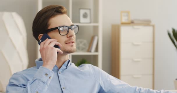 Közelkép a fiatal vonzó férfi szemüvegben válaszol a hívást, és beszél telefonon a szép nappaliban. Portré felvétel. Belül. — Stock videók