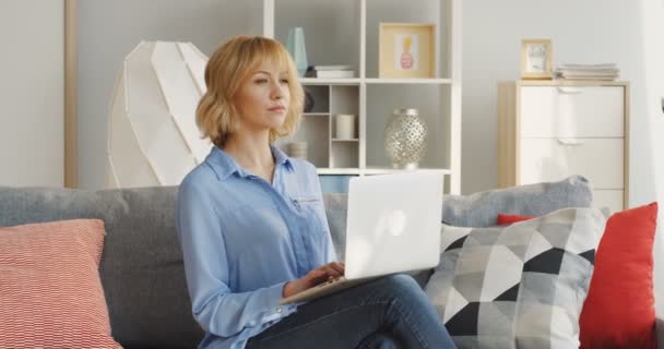 Blonde blanche belle femme qui travaille sur l'ordinateur portable à la maison et de penser tout en étant assis sur le canapé confortable. Intérieur — Video