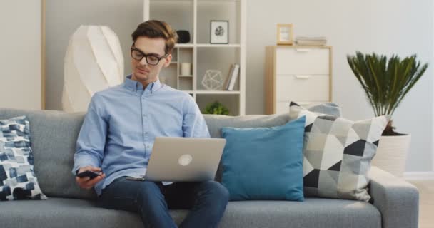 Kaukasischer Jungunternehmer, der den Anruf entgegennimmt, während er zu Hause auf der Couch am Laptop arbeitet. Innenräume — Stockvideo