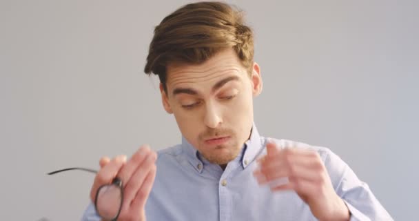 Portrait of the young caucasian tired man taking off glasses and worring a lot in front of the camera on the gray wall background. Close up. Indoor — Stock Video