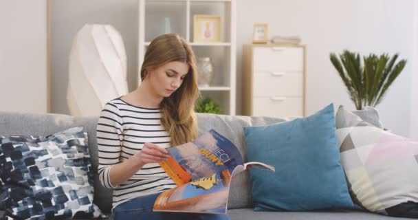 Retrato filmado da jovem mulher bonita sentada no sofá na sala de estar e virando páginas na revista das mulheres. Interior — Vídeo de Stock