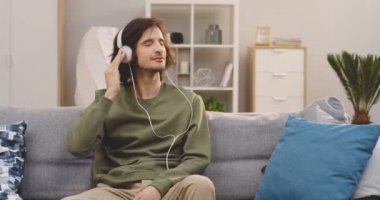 Genç adam elinde akıllı bir telefonla kanepede oturuyor ve kulaklıkla müzik dinliyor. - Evde. Kapalı