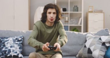 Beyaz tenli, uzun saçlı, elinde joystick ile video oyunları oynayan çekici bir adam oturma odasında yastıklarla gri koltukta oturuyor. İçeride.