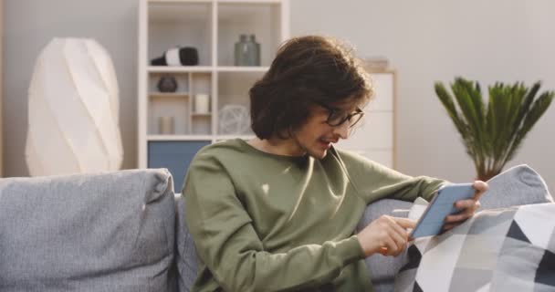 Homem bonito jovem com um longo cabelo rolando e gravando no dispositivo tablet, assistindo algo e rindo no sofá em casa. Interior — Vídeo de Stock