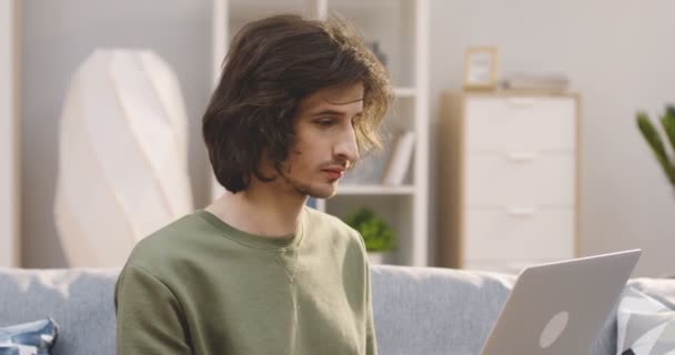 Primer plano del joven con el pelo largo trabajando y escribiendo en el ordenador portátil en la sala de estar en casa. Retrato. De interior — Vídeos de Stock