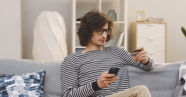 Joven estudiante de cabello largo y gafas que compra online en el smartphone e introduce los datos de la tarjeta de crédito en el sofá de la sala de estar. Adentro. — Vídeos de Stock