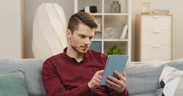 Fiatal vonzó fehér férfi a piros sötét sh ül a szürke kanapén, és használja a tabletta eszköz, görgetés és néz valamit. Otthon. Belül. — Stock videók