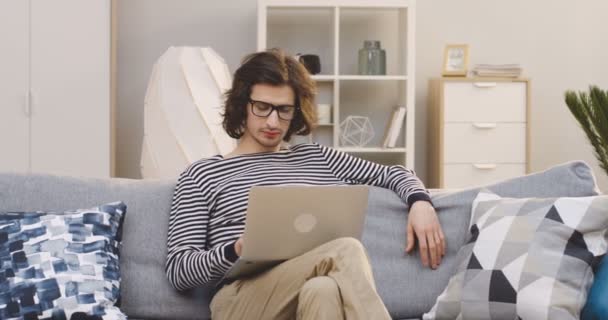 Молодий студент-чоловік в окулярах працює на портативному комп'ютері вдома і думає, сидячи на затишному дивані. індорів — стокове відео