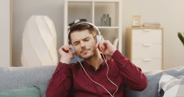 Jovem bonitão de camisa vermelha escura sentado no sofá cinza na sala de estar ouvindo a música nos fones de ouvido e se movendo no ritmo. Em casa. Para dentro — Vídeo de Stock