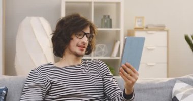 Gözlüklü beyaz erkek öğrenci evdeki kanepede otururken tablet bilgisayarında görüntülü sohbet ediyor. Kapalı