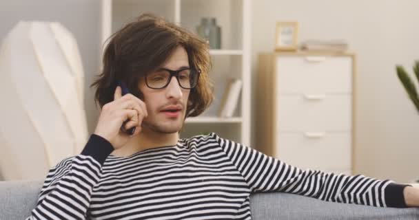 안경을 쓰고 줄 무늬 스웨터를 입고 소파에 앉아 전화를 걸고 대화를 나누고 있는 코카서스 청년. 초상화. 실내에서 — 비디오
