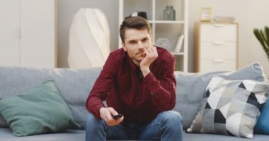 Uzaktan kumandalı genç ve yakışıklı bir adam kanepede oturup televizyon seyrediyor ve kanal değiştiriyor. - Evde. Kapalı