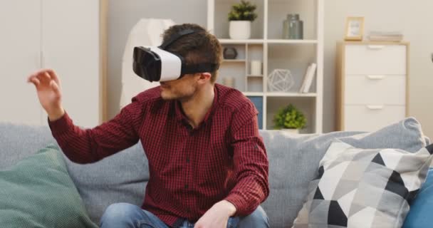 年轻英俊的高加索男子头戴VR耳机，头戴VR眼镜，沙发上的枕头在客厅里的肖像照片。室内 — 图库视频影像