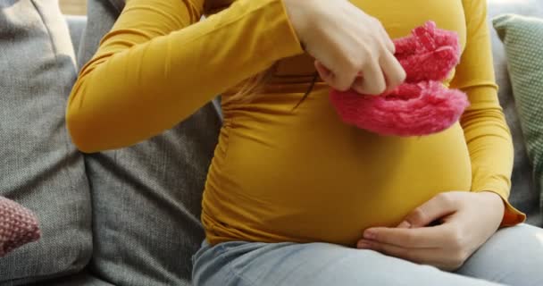 Primer plano de la barriga de las mujeres embarazadas y las manos con los niños pequeños zapatos de color rosa en frente de ella. Sofá gris. En interiores — Vídeos de Stock