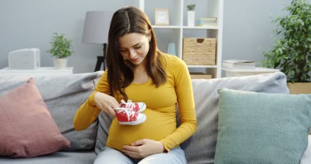 Mujer embarazada caucásica jugando con zapatos pequeños rojos y blancos y pensando en su hijo en el sofá de la sala de estar. Adentro. — Vídeos de Stock