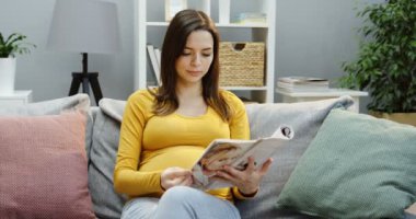 Güzel hamile kadın kanepede yastıklarla oturuyor ve rahat oturma odasında dergi okuyor. Kapalı