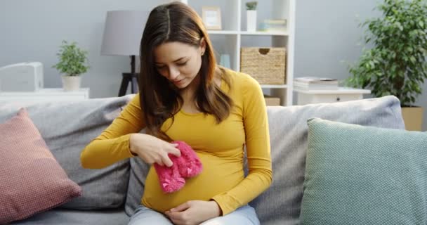 Attraktive schwangere Frau, die mit kleinen rosa Schuhen spielt, während sie auf dem Sofa im Wohnzimmer sitzt. Porträtaufnahme. Innenräume — Stockvideo