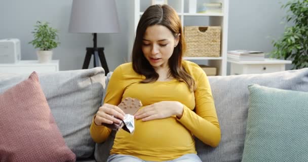 Bella donna incinta mangiare una tavoletta di cioccolato con noci mentre seduto sul divano grigio con cuscini nell'accogliente soggiorno moderno. Dentro. — Video Stock