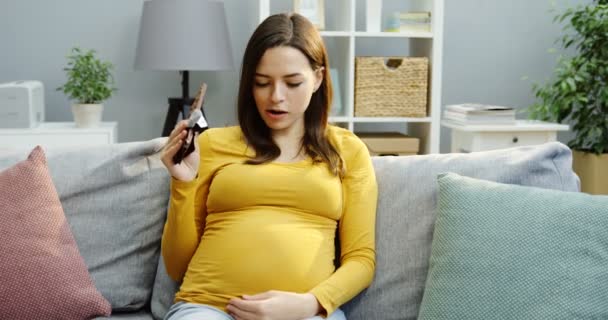Charmante femme enceinte assise sur le canapé gris, mordant et mâchant du chocolat dans la chambre confortable. Manger une barre de chocolat. À l'intérieur — Video